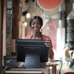 Positive young woman in uniform smiling while standing at counter desk in cafe